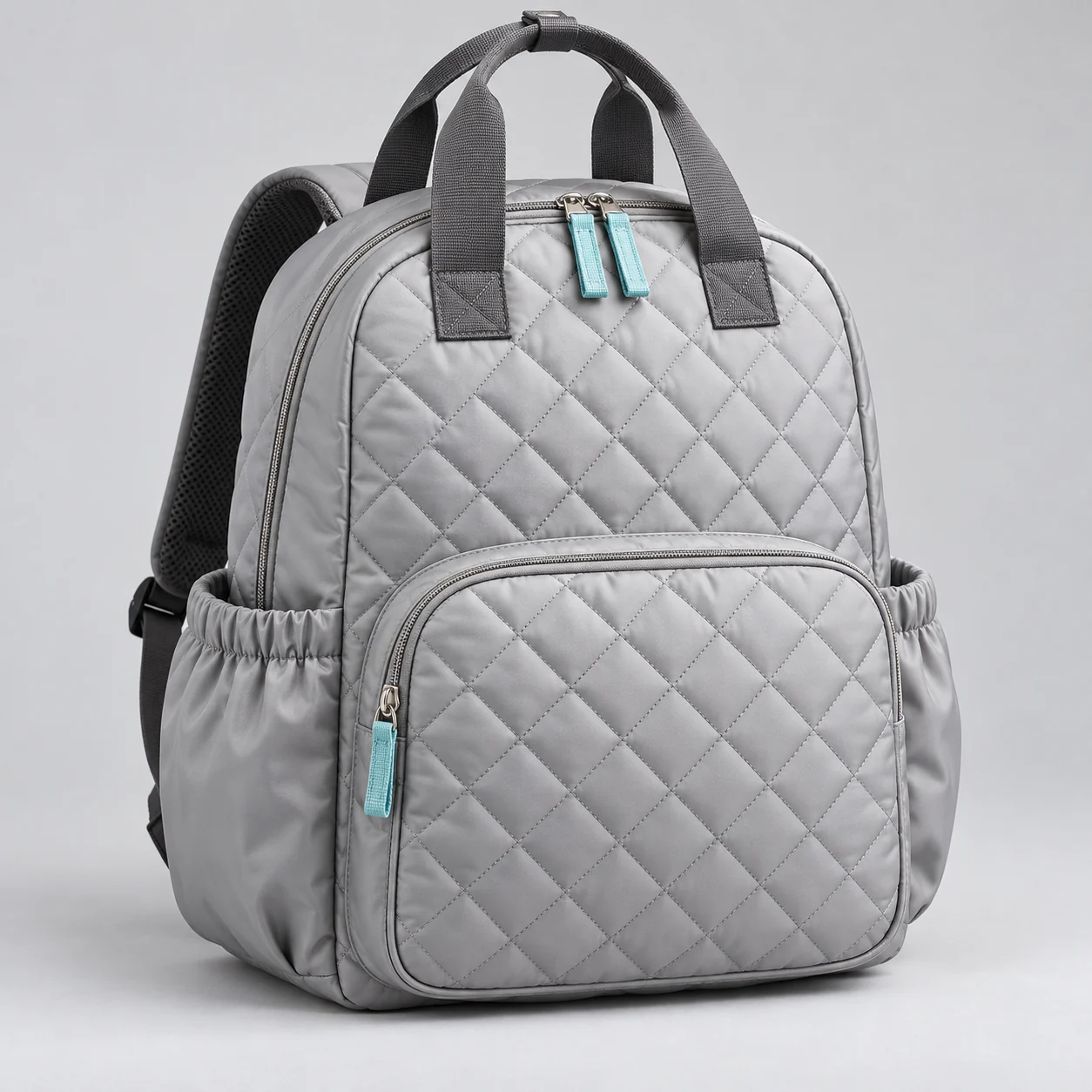 Silver-gray quilted changing backpack with dark handles, side pockets and aqua zipper pulls on a pale background.